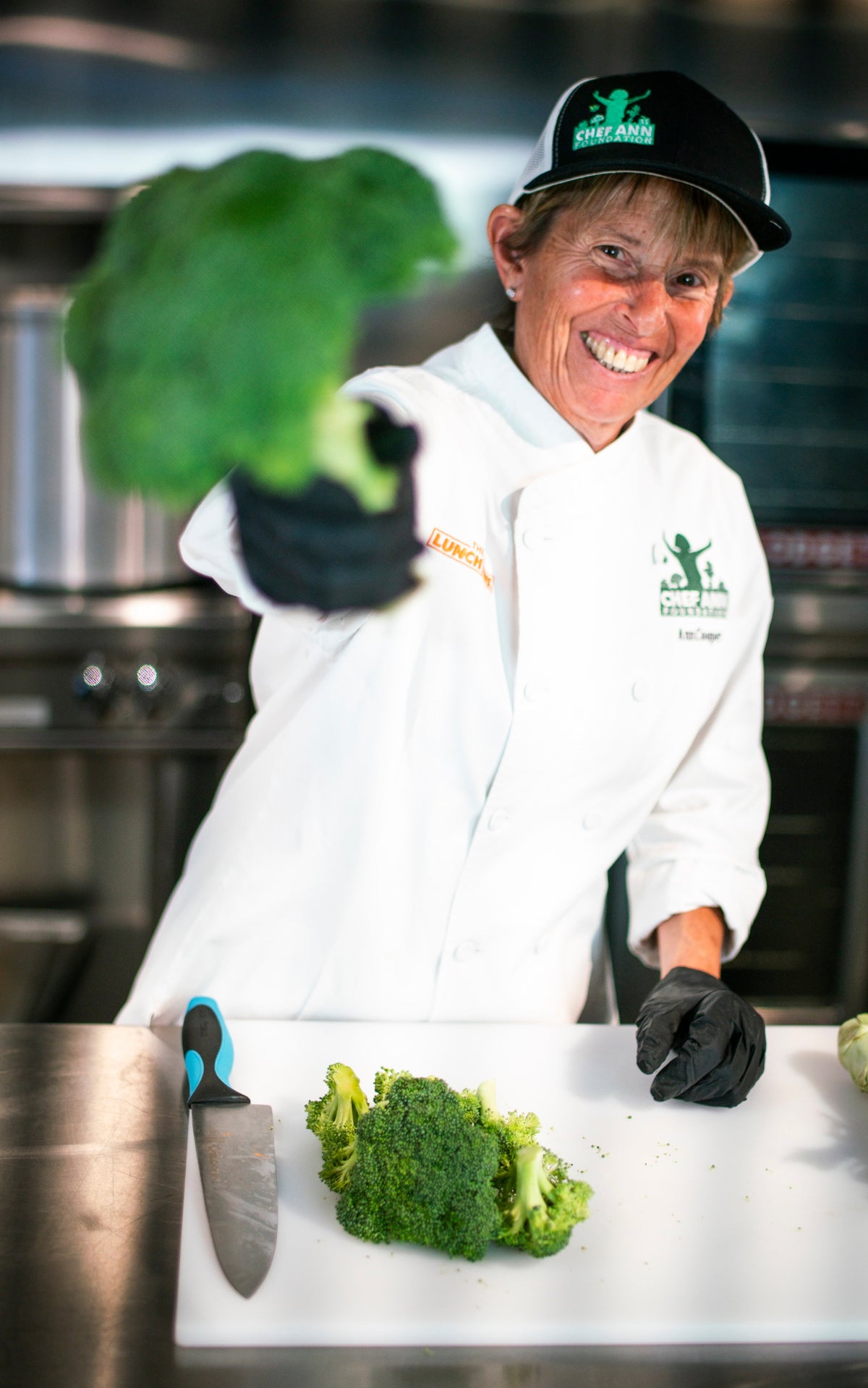 A smiling Chef Ann Cooper in a white chef coat and black cap holds a large piece of broccoli toward the camera in a commercial kitchen setting, with a cutting board, knife, and smaller broccoli pieces on the counter.
